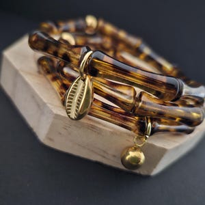May include: A stack of brown and tan bamboo-style bracelets with gold-colored charms. One charm is a small shell shape, and another is a round ball. The bracelets are displayed on a small, light-colored wooden block.