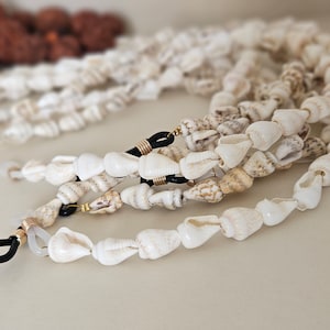 Peut inclure: Chaîne de lunettes en coquillages blancs avec des accents dorés et noirs. La chaîne est composée de plusieurs brins de coquillages.