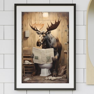May include: Framed art print featuring a moose reading a newspaper while seated on a toilet. The artwork has a humorous theme, with the moose in a rustic bathroom setting. The print is in a black frame and is displayed on a white tiled wall.