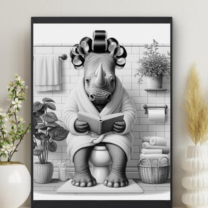 May include: Monochrome framed print depicting a rhino in a bathrobe and hair curlers, reading a book on a toilet. The bathroom scene includes a towel rail, potted plants, and a basket of folded towels.