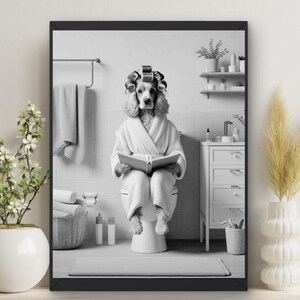 May include: A black and white photo of a dog wearing a white bathrobe and sitting on a toilet. The dog has rollers in its hair and is reading a book. The photo is framed in a black frame.