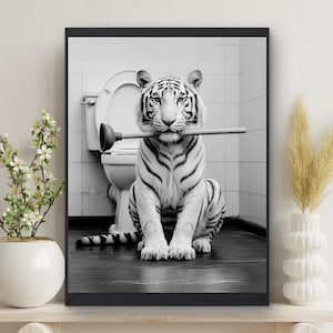 May include: A black and white photograph of a white tiger sitting in a bathroom with a plunger in its mouth. The tiger is looking directly at the camera.