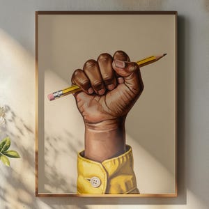 May include: A framed art print featuring a dark-skinned hand clenched around a yellow pencil. The hand is wearing a yellow shirt with a buttoned cuff. The background is a neutral beige.