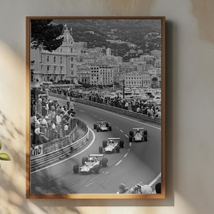 Może przedstawiać: Oprawione czarno-białe zdjęcie zabytkowego wyścigu Formuły 1. Obraz przedstawia samochody wyścigowe na torze z widzami i budynkami w tle. Biały wazon z kwiatami, mały dzbanek i filiżanka kawy znajdują się na drewnianej powierzchni.