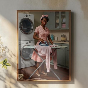May include: A framed print depicts a woman ironing clothes in a vintage kitchen. She wears a pink dress and headband, using a blue iron on a pink garment. A washing machine is visible behind her. A coffee cup and vase are in the foreground.
