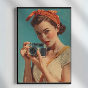 Puede incluir: Impresión enmarcada de estilo vintage que presenta a una mujer sosteniendo una cámara plateada y negra. Lleva una blusa blanca, un pañuelo naranja y lápiz labial rojo sobre un fondo azul claro. La impresión está en un marco negro.