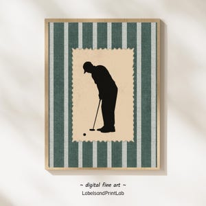 May include: A framed digital art print showing a black silhouette of a golfer putting. The artwork is set against a green and white striped background, with the golfer image inside a postage stamp-like border. The frame is a light wood colour.