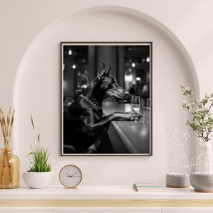 Peut inclure: Photographie en noir et blanc d'un chien Dobermann assis &agrave; un comptoir de bar avec un verre de vin. Le chien porte un collier argent&eacute; et regarde le verre de vin.