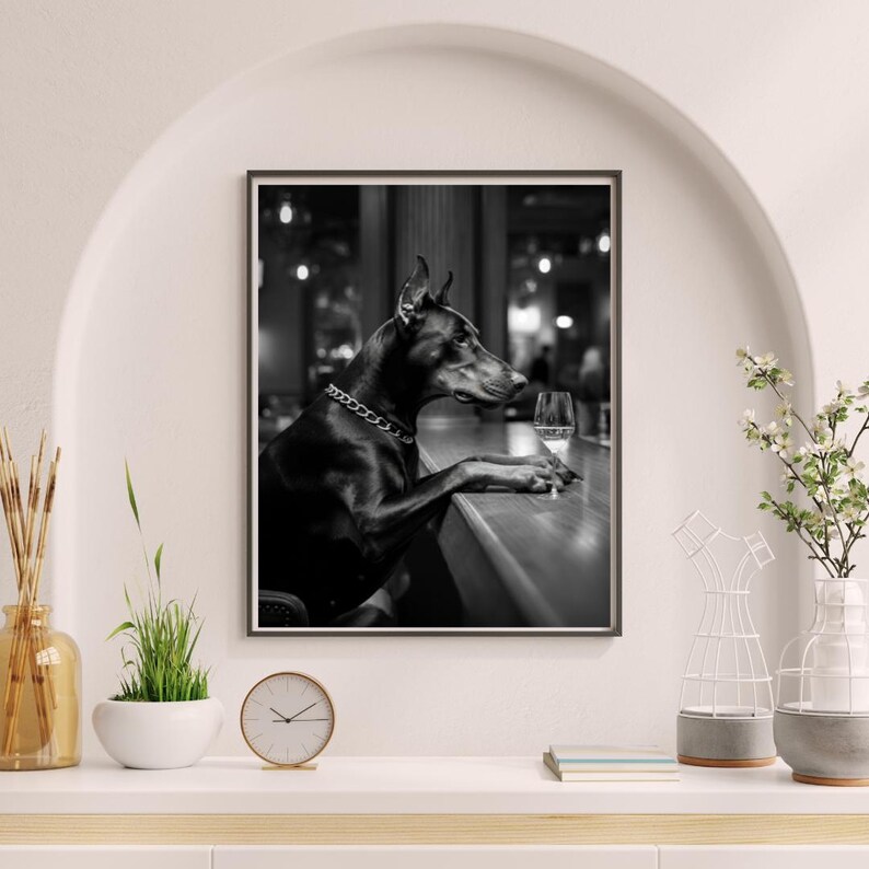 Peut inclure: Photographie en noir et blanc d'un chien Dobermann assis &agrave; un comptoir de bar avec un verre de vin. Le chien porte un collier argent&eacute; et regarde le verre de vin.