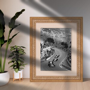 Puede incluir: Fotograf&iacute;a en blanco y negro enmarcada de coches de carreras en una pista urbana. La imagen est&aacute; dentro de un marco dorado adornado con un passe-partout blanco. La foto captura la emoci&oacute;n de una carrera, con coches en una carretera sinuosa.