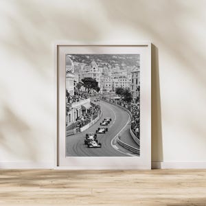 Puede incluir: Impresi&oacute;n enmarcada en blanco y negro de una carrera de F&oacute;rmula 1 en M&oacute;naco. La imagen captura coches de carreras en una pista sinuosa, bordeada de espectadores y edificios. La fotograf&iacute;a se presenta en un marco blanco.