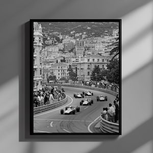 Może przedstawiać: Czarno-biała oprawiona grafika przedstawiająca zabytkowy wyścig Formuły 1. Kilka bolidów wyścigowych z otwartymi kołami pędzi po krętym torze, otoczonym przez widzów i bariery. Tło przedstawia krajobraz miejski z budynkami i wzgórzami.