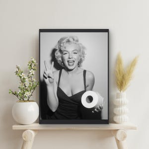 May include: Black and white framed print featuring Marilyn Monroe holding a roll of toilet paper and making a peace sign. The print is displayed on a wooden shelf with decorative plants and a vase.