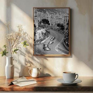 Puede incluir: Fotograf&iacute;a en blanco y negro enmarcada de una carrera de F&oacute;rmula 1 en M&oacute;naco. La imagen captura coches de carreras en una pista sinuosa, bordeada de espectadores y edificios. La fotograf&iacute;a se muestra sobre una superficie de madera, con un jarr&oacute;n, una lechera y una taza de caf&eacute;.