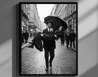Può includere: Stampa incorniciata in bianco e nero di un uomo che porta una donna per una strada acciottolata. Entrambi sono vestiti di scuro e l'uomo tiene un ombrello nero. La scena evoca un'atmosfera vintage e romantica.