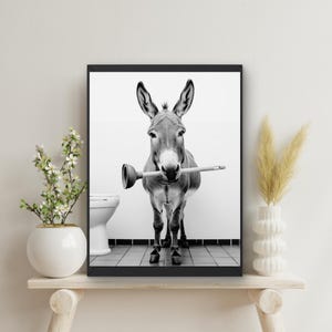 Op de afbeelding: Een zwart-witfoto van een ezel die een zuiger in zijn mond houdt. De ezel staat in een badkamer met een toilet op de achtergrond.
