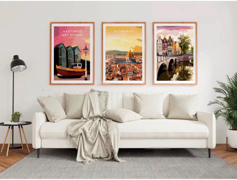 a living room with a white sofa and a potted plant. Above the sofa, there are three framed posters depicting different cities: Hasting's Net Shops in London, Florence in Italy, and Amsterdam in the Netherlands.