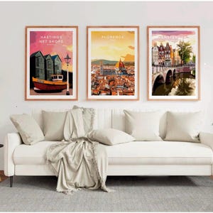 a living room with a white sofa and a potted plant. Above the sofa, there are three framed posters depicting different cities: Hasting's Net Shops in London, Florence in Italy, and Amsterdam in the Netherlands.