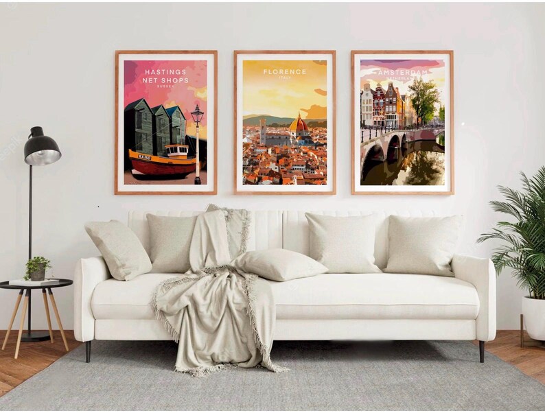 a living room with a white sofa and a potted plant. Above the sofa, there are three framed posters depicting different cities: Hasting's Net Shops in London, Florence in Italy, and Amsterdam in the Netherlands.