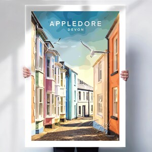 May include: A colorful art print of Appledore, Devon, featuring a street scene with pastel-colored buildings and seagulls in flight. The print has the text "Appledore Devon" at the top. The artwork is held by a person.