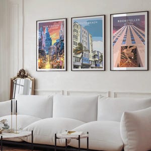 a living room with a white sofa and a coffee table. Above the sofa, there are three framed posters of different cities, including New Orleans, Miami Beach, and Rockefeller Center, which are displayed on the wall.