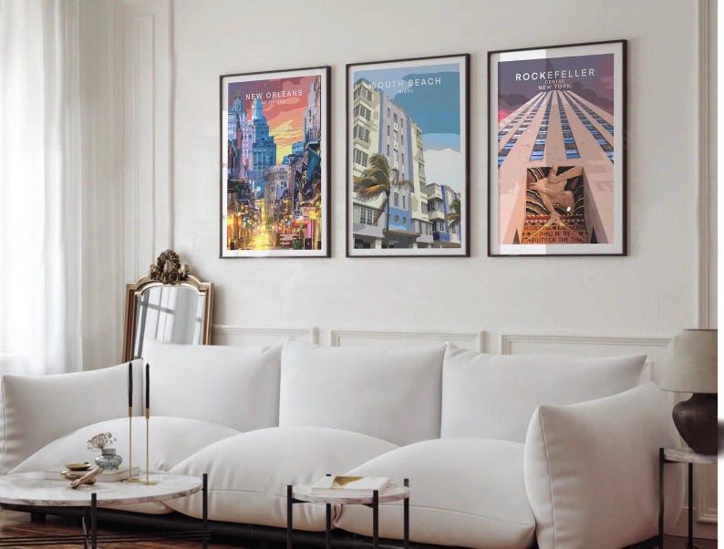 a living room with a white sofa and a coffee table. Above the sofa, there are three framed posters of different cities, including New Orleans, Miami Beach, and Rockefeller Center, which are displayed on the wall.