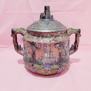 May include: A ceramic pitcher with a lid, featuring a detailed design of a castle and grapevines. The pitcher has two handles shaped like dragon heads and is decorated with green, brown, and purple accents.