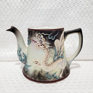 Peut inclure: Une théière en céramique avec un motif de dragon. La théière présente un dragon blanc avec des détails complexes, sur un fond dégradé de noir, gris et blanc. La poignée et le bec sont noirs et blancs, avec des accents bleus.