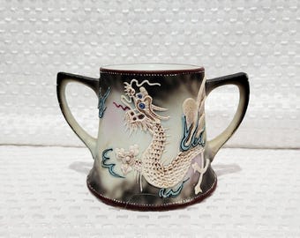 Op de afbeelding: Een decoratieve keramische mok met twee handvatten, met een gedetailleerd reliëf van een draak. De mok heeft een gradiënt achtergrond van zwart en wit, met blauwe en rode accenten. De rand en handvatten zijn donkerbruin.