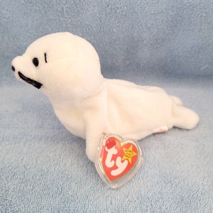 May include: White plush seal beanie baby toy with black eyes and a black moustache. The toy has a red heart-shaped tag with the words "Ty" and "Beanie Baby" printed on it.