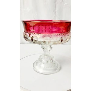 Puede incluir: Un cuenco de pedestal de vidrio vintage con una base transparente y una parte superior roja, transparente y blanca. El cuenco tiene un borde festoneado y un diseño texturizado.