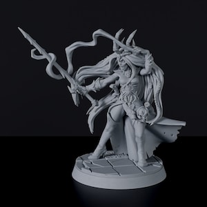 May include: A gray 3D printed miniature of a female figure with horns, long hair, and a flowing robe. She holds a spear in her right hand and a small object in her left hand. The figure is standing on a round base.