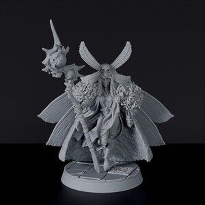 May include: A gray, detailed miniature figurine of a female figure with a skull headdress, feathered wings, and a staff with a skull and floral accents. The figure is standing on a circular base.