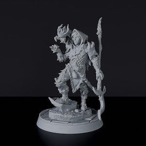 May include: A gray, detailed miniature figurine of a hooded figure with a horned creature on their shoulder, holding a staff with a curved blade. The figure is standing on a circular base with a textured surface.