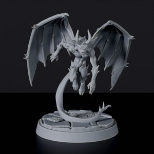 Op de afbeelding: Een grijs, gedetailleerd miniatuurbeeldje van een demon met vleugels, een lange staart en scherpe klauwen. De demon is in de lucht geposeerd, met zijn vleugels wijd uitgespreid. Het beeldje staat op een ronde basis.