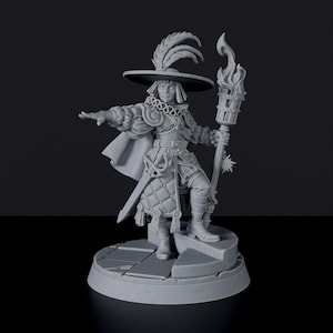 May include: A gray miniature figure of a person in historical attire, holding a torch. The figure wears a hat with feathers, a cape, and a sword. The figure stands on a circular base with a step.