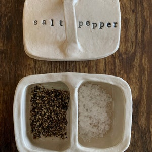 以下が含まれることがあります： 塩とコショウの入れ物セット。白いセラミック製で、それぞれのスパイスを入れるための仕切りがあります。蓋には「塩」と「コショウ」の文字が刻まれています。