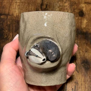 May include: A ceramic mug with a 3D badger design. The badger is brown and white with black eyes and a black nose. The mug is a light brown color with a textured surface.
