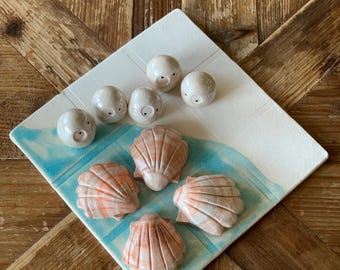 May include: A white square ceramic plate with a blue and white wave design. The plate holds six small, round, off-white sculptures with tiny black eyes, and five pink and white seashell sculptures.