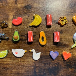 Peut inclure: Une collection de 17 breloques en céramique, chacune avec un design unique. Les breloques comprennent une banane, un cœur, un poisson, une papaye, un citron vert, un canard, un avocat, un chat, une tasse, une oie, et plus encore. Les breloques sont toutes peintes à la main et présentent une variété de couleurs.