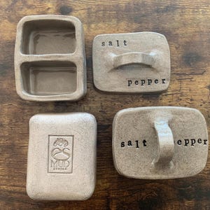 Puede incluir: Un juego de cuatro saleros y pimenteros de cerámica. Dos de los saleros son rectangulares con una manija y las palabras "sal" y "pimienta" inscritas en ellos. Los otros dos saleros son cuadrados, uno con un compartimento dividido y el otro con un logotipo que dice "es mud zürich".