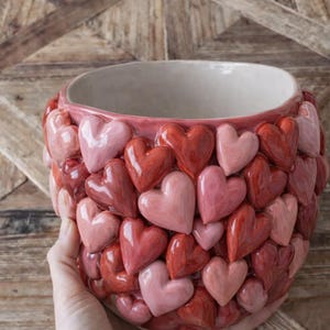 Puede incluir: Una maceta de cerámica con diseño de corazón. La maceta presenta una mezcla de corazones rojos y rosas. El interior es de color beige claro. La maceta está sostenida por una mano.