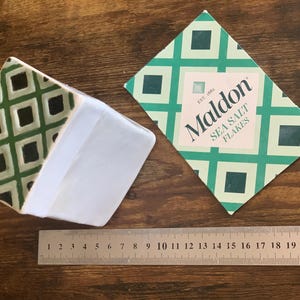 Può includere: Vista dall'alto di una confezione di Maldon Sea Salt Flakes e di un sottobicchiere fantasia. La confezione è verde e bianca con il nome del marchio. Un righello è posto sotto. Il sottobicchiere ha un motivo geometrico verde e nero.