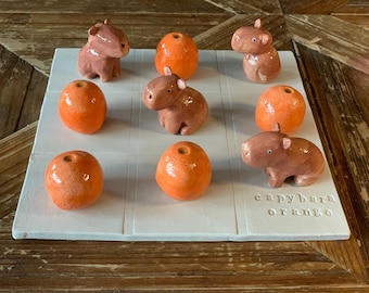 Può includere: Un gioco in ceramica del tris con sfere arancioni e statuette di capibara marroni. Il tabellone bianco ha un motivo a griglia. Il testo "capybara orange" è stampato sul tabellone.