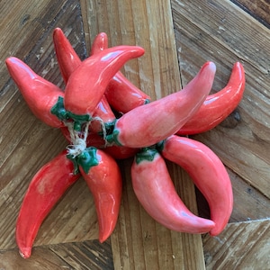 Könnte beinhalten: Ein Cluster aus roten Keramik-Chilischoten, die mit Bindfaden zusammengebunden sind. Die Paprika haben grüne Stiele und eine glänzende Oberfläche. Die Anordnung befindet sich auf einem Holzgrund.