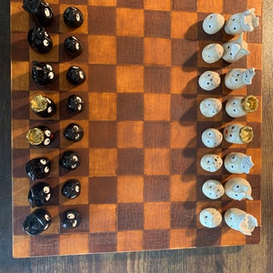 Puede incluir: Un tablero de ajedrez de madera con piezas de ajedrez de cerámica negra y blanca. Las piezas negras tienen ojos saltones y las piezas blancas tienen un diseño de pájaro. El tablero está configurado para una partida de ajedrez.