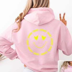 Könnte beinhalten: Hellrosa Kapuzenpullover mit Kängurutasche und Kapuze. Auf der Rückseite befindet sich ein gelbes Smiley-Gesicht mit Herzaugen und dem Schriftzug "MORE AMORE" in einem Kreis.