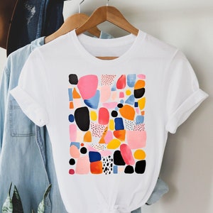 Könnte beinhalten: Weißes T-Shirt mit einem farbenfrohen abstrakten Design mit rosa, blauen, gelben, orangen und schwarzen Formen.