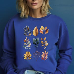 Könnte beinhalten: Ein königsblaues Langarm-Sweatshirt mit einem zentralen Grafikdruck aus neun verschiedenen botanischen Illustrationen. Das Design zeigt verschiedene Blatt- und Zweigmotive in Orange-, Gelb-, Braun- und gedämpften Blautönen, die in einem Raster angeordnet sind.