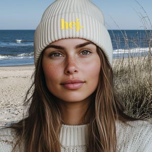 May include: A cream-coloured ribbed knit beanie featuring the word "hej" in yellow. The hat is worn by a person with long brown hair and a cream-coloured sweater. The background shows a beach scene with the sea and tall grass.